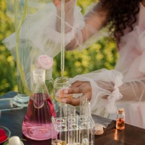 A woman with curly hair mixing perfumes outdoors with glassware and pipette.