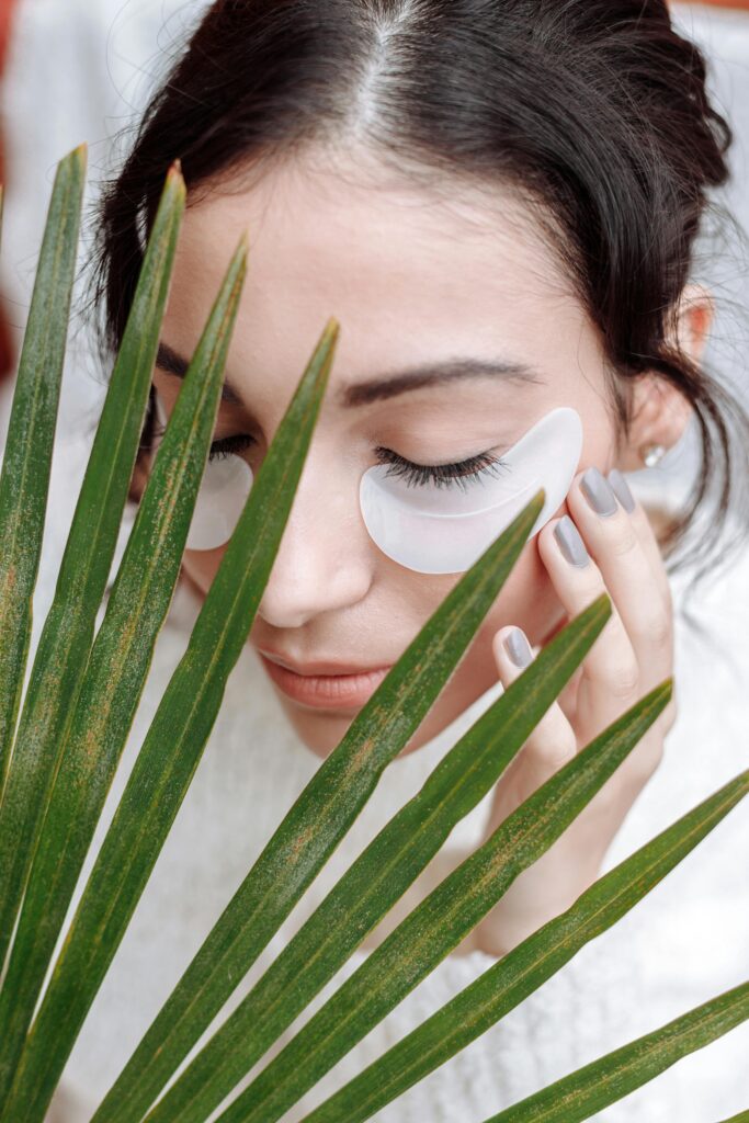 Close-up of a woman applying skincare under eye masks, surrounded by green leaves.